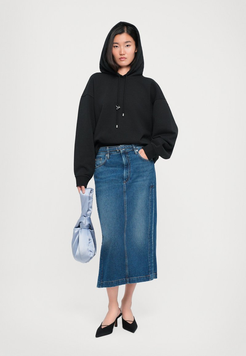 Haut à capuche noir surdimensionné, jupe midi en denim bleu, talons noirs à bout pointu, tenant un sac en tissu bleu clair avec une anse nouée. Design simple et décontracté.