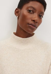 Beige hochgeschneiderter Strickpullover mit weicher Textur und geripptem Kragen. Das Model trägt einen schlichten silbernen Creolenohrring.