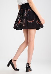 Žena v černé květované sukni a černých lakových botách na blokovém podpatku kráčí před jednobarevným pozadím.