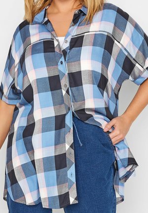 Chemise ample à manches courtes à carreaux bleu, noir et blanc portée sur un top blanc avec un jean bleu, une main dans la poche.