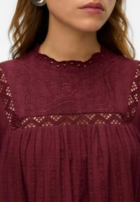 Burgundy blus med intrikata broderier och spetsdetaljer, med en vågig halsringning och rynkad struktur genom hela plagget.
