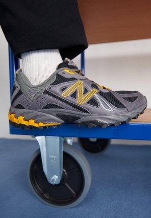 Stopa ubrana w szaro-żółte buty sportowe New Balance z białą skarpetką, spoczywająca na niebieskim metalowym kole wózka w pomieszczeniu.