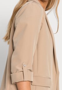 Beige blazer with a soft texture, featuring rolled sleeves with a pearl button accent and a structured design.