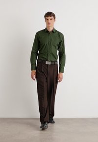 Cămașă verde cu nasturi, cu o textură netedă, purtată cu pantaloni maro. Asortată cu o curea texturată neagră și pantofi negri. Design simplu.