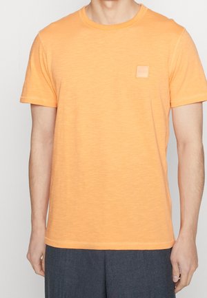 Man die een oranje T-shirt met korte mouwen en ronde hals draagt, met een klein vierkant embleem op de borst en donkergrijze broek, staand tegen een effen achtergrond.
