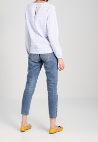 Helle blau gestreifte Bluse mit langen Ärmeln und einem Rückenausschnitt, kombiniert mit blauen Jeans und gelben Ballerinas, die einen lässigen Look präsentieren.
