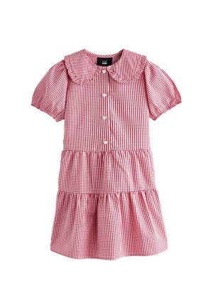 Smiješeća mlada djevojka u crveno-bijeloj kariranoj haljini s nabranim rukavima i škarastim ovratnikom, prekriženih ruku, gleda blago prema gore.