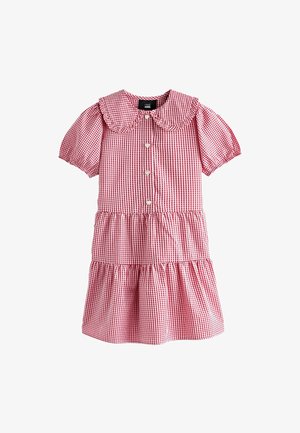 Lachend jong meisje in een rood-wit geruite jurk met pofmouwen en een geschulpte kraag, armen over elkaar, kijkt iets omhoog.