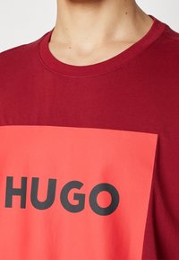 Camiseta burdeos con un gran gráfico rectangular rojo con el texto "HUGO" en negro. Cuello redondo y mangas cortas; textura de tela suave.