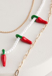Red chili pepper charms with green accents on a white pearl bead necklace, paired with a gold link chain. Textured and colorful design.