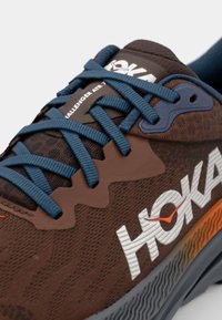 Hnědá sportovní obuv s modrými šňůrkami, texturovaným svrškem a velkým logem „HOKA“. Obsahuje gradientní design podrážky v oranžové a šedé barvě.