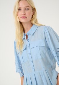 Svetlo modra srajčna obleka z vezenimi cvetličnimi detajli in okrasnimi luknjami, ki ima kratke napihnjene rokave in dva žepa na prsih.