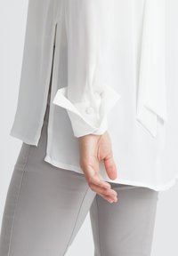 Witte sheer blouse met lange mouwen, knoopmanchetten en zijsplitten, gecombineerd met lichtgrijze, aansluitende broek. Soepele, lichte stof.