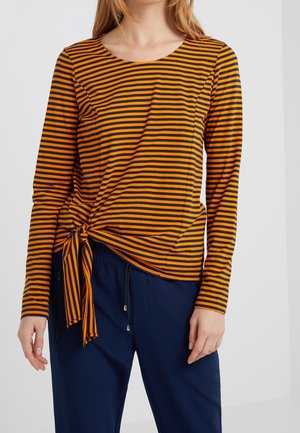 Vrouw die een oranje en marineblauw gestreept shirt met lange mouwen en een voorstrikdetail draagt, samen met marineblauwe broek met trekkoord, staand tegen een witte achtergrond.