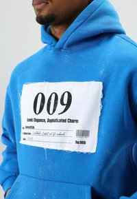 Sweat à capuche en coton bleu avec une grande étiquette blanche à texte noir, un patch texturé et une poche avant. Les détails de l'étiquette incluent "009" et "Élégance Iconique."