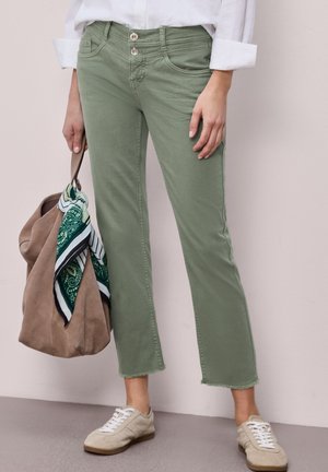 Persoon die olijfgroene korte broek met rafelige zomen, witte sneakers en een opgerolde witte blouse draagt, en een beige tas vasthoudt met een groene sjaal met patroon.