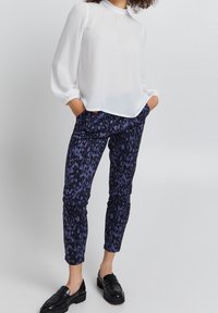 Blusa bianca a maniche lunghe con scollo arricciato; abbinata a pantaloni attillati blu navy con un motivo a leopardo viola testurizzato. Mocassini neri completano il look.