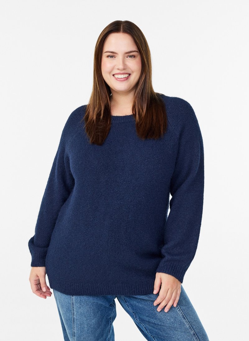 Donna sorridente con lunghi capelli castani che indossa un maglione blu navy e jeans blu, in piedi davanti a uno sfondo bianco semplice.