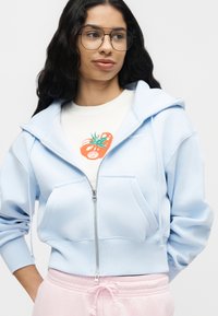 Νέα γυναίκα με μακριά σκούρα μαλλιά και γυαλιά φοράει ανοιχτό μπλε φούτερ με φερμουάρ, λευκό μπλουζάκι με γραφικό τομάτας και ανοιχτό ροζ παντελόνι.