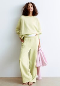 Hellgrüner Sweatshirt mit oversized Schnitt, kombiniert mit passenden weit geschnittenen Hosen; die Textur erscheint weich. Ein pinkes Oberteil hängt über einem Arm.