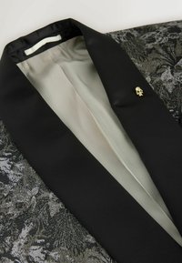 Black blazer featuring a textured fabric with a floral pattern. Notable details include a gold skull pin and a smooth inner lining.