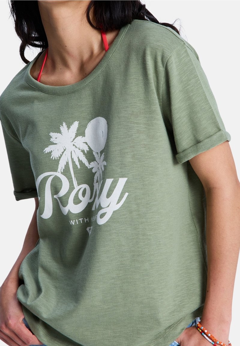Femme portant un t-shirt vert Roxy avec un motif de palmier blanc et des bretelles de bikini rouges visibles, mains reposant près des hanches.