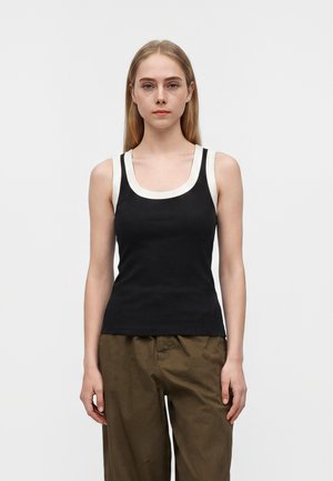 Jeune femme aux cheveux blonds raides portant un débardeur noir avec une bordure blanche et un pantalon marron ample, debout devant un fond uni.