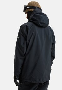 Schwarze wasserdichte Jacke mit Kapuze, aus strukturiertem Stoff, verstellbaren Manschetten und einem Logo-Patch am Ärmel.