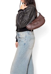 Borsa a tracolla in pelle marrone con chiusura a zip, forma curva e sottili dettagli metallici, indossata sopra una giacca bomber nera e jeans chiari.