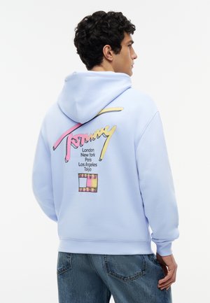 Junger Mann trägt einen hellblauen Kapuzenpullover mit buntem "Tommy"-Schriftzug und Stadtnamen auf dem Rücken, kombiniert mit blauen Jeans.