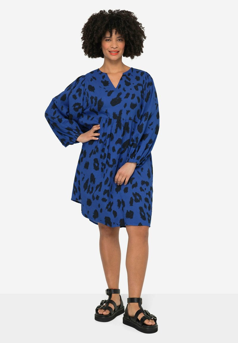 Robe bleue avec motif léopard noir, décolleté en V, manches longues ballons, taille cintrée et longueur au-dessus du genou. Portée avec des sandales noires à lanières.