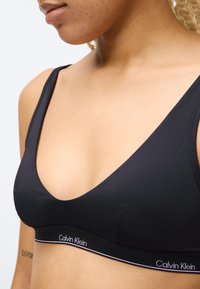 Κοντινό πλάνο γυναίκας που φοράει ένα μαύρο αθλητικό σουτιέν Calvin Klein με λεπτές τιράντες, δείχνοντας μέρος του λαιμού, του ώμου και του άνω στήθους της.