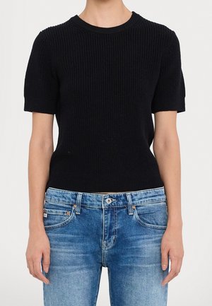 Personne portant un pull en maille noir à manches courtes et un jean en denim bleu, les bras détendus le long du corps, sur un fond uni.