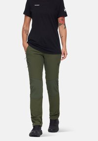 Groene outdoorbroek met een slanke pasvorm, gemaakt van duurzaam materiaal. Voorzien van kniebeschermers en zijzakken, gecombineerd met een zwart T-shirt en laarzen.