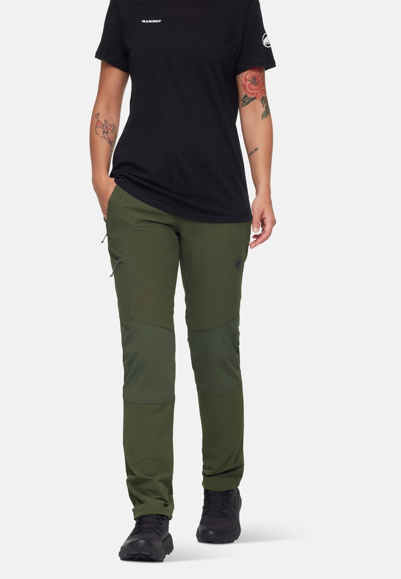 Groene outdoorbroek met een slanke pasvorm, gemaakt van duurzaam materiaal. Voorzien van kniebeschermers en zijzakken, gecombineerd met een zwart T-shirt en laarzen.