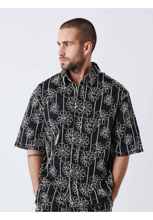 Homme aux cheveux courts et barbe, portant une chemise noire à manches courtes avec une broderie florale blanche, mains dans les poches, regardant vers la gauche sur un fond uni.