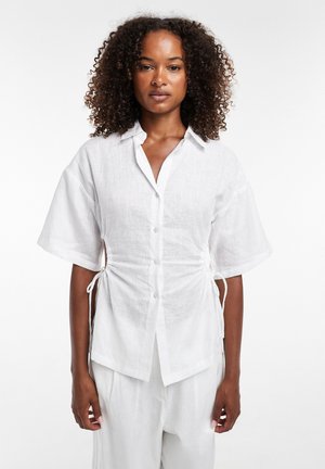 Kvinde med krøllet hår iført en hvid kortærmet skjorte med sidelukninger og matchende hvide bukser mod en ensfarvet baggrund.