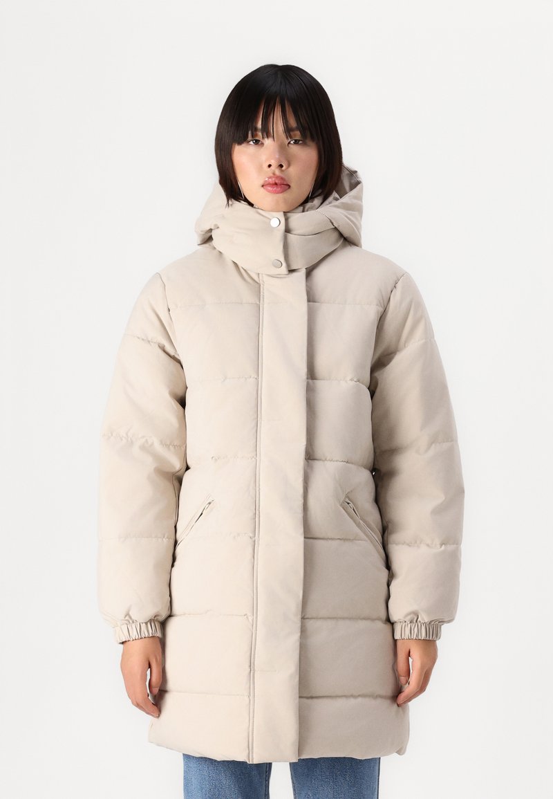 Manteau doudoune beige avec un col montant et une capuche ajustable. Présente des poignets élastiques et des poches latérales. Design matelassé horizontal sur l'ensemble.