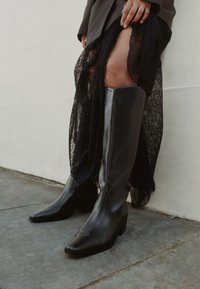 Bottes hautes en cuir noir jusqu'aux genoux avec un bout pointu et un talon bloc, présentant une texture lisse et un design minimaliste, associées à une jupe en dentelle noire transparente.