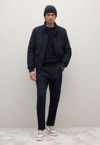 Marineblaue Wolljacke mit Knopfleiste, kombiniert mit einem dunklen Pullover und maßgeschneiderten marineblauen Hosen. Beigefarbene und weiße Sneaker vervollständigen den Look.