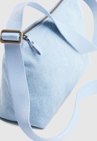 Φωτεινή γαλάζια τζιν τσαντάκι ώμου με ρυθμιζόμενο λουρί και φερμουάρ, σε απλό ανοιχτό φόντο.