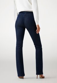 Jean flare en denim bleu foncé avec une taille mi-haute, dotés de poches arrière et de coutures détaillées, portés avec un pull blanc et des talons.