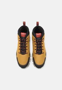 Botas de senderismo amarillas con acentos negros, superficie texturada, cordones rojos y negros, y lengüetas con un logo en el talón.