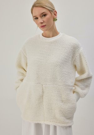 Femme portant un pull ample, crème, duveteux avec des poches et des boucles d'oreilles en forme de feuilles vertes, debout sur un fond uni.