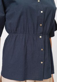 Camicia blu navy a maniche corte, vita arricciata e dettagli dorati lungo la parte anteriore, realizzata in un tessuto leggero e testurizzato.