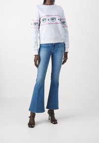 Vit sweatshirt med ett grafiskt band av ögon och stjärnor, matchad med utsvängda blå jeans och svarta klacksandaler.