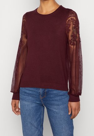 Femme portant un pull bordeaux avec des manches en dentelle florale transparente et un jean bleu, debout devant un fond clair uni.