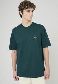 T-shirt en coton vert foncé à manches courtes, encolure ronde et texte imprimé blanc "Meet me Playground" sur la poitrine gauche.