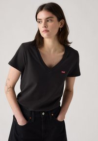 T-shirt noir en coton avec un col en V et des manches courtes, arborant un petit logo rouge Levi's sur la poitrine. Porté avec un jean noir taille haute.