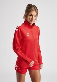 Rode sportjack met een hoge kraag, ritssluiting en zijzakken. Heeft witte chevronaccenten op de mouwen en branding op de borst.